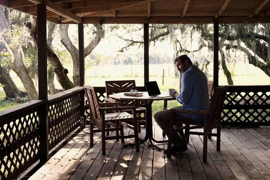 Jeremy working at a table on a ranch porch beneath live oaks